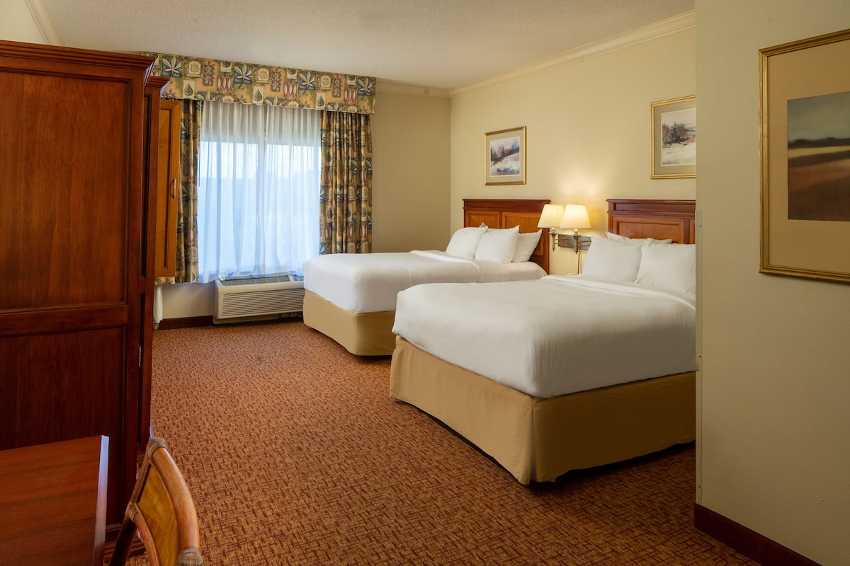 A hotel room with two beds, a wooden wardrobe, a window with curtains, and a framed picture on the wall.