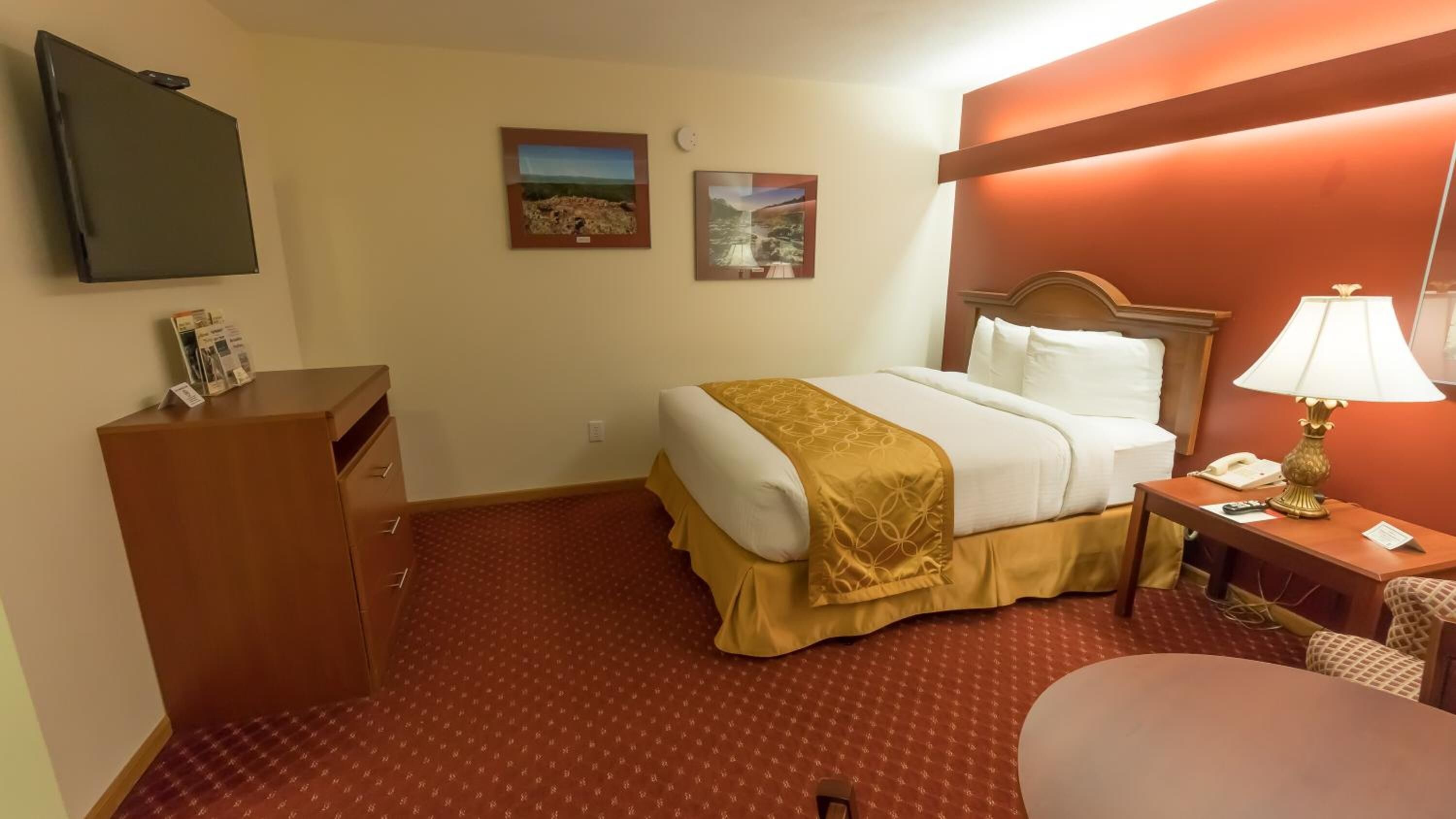 A hotel room with a bed, a desk, a chair, a TV, and two framed pictures on the wall.