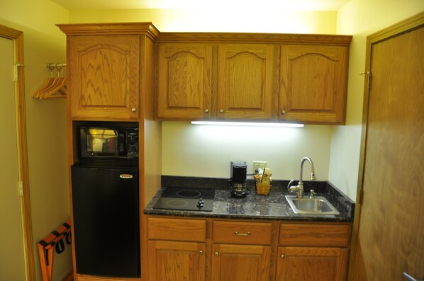 Cocina compacta con armarios de madera, un refrigerador negro, microondas integrado, cafetera y un lavamanos.