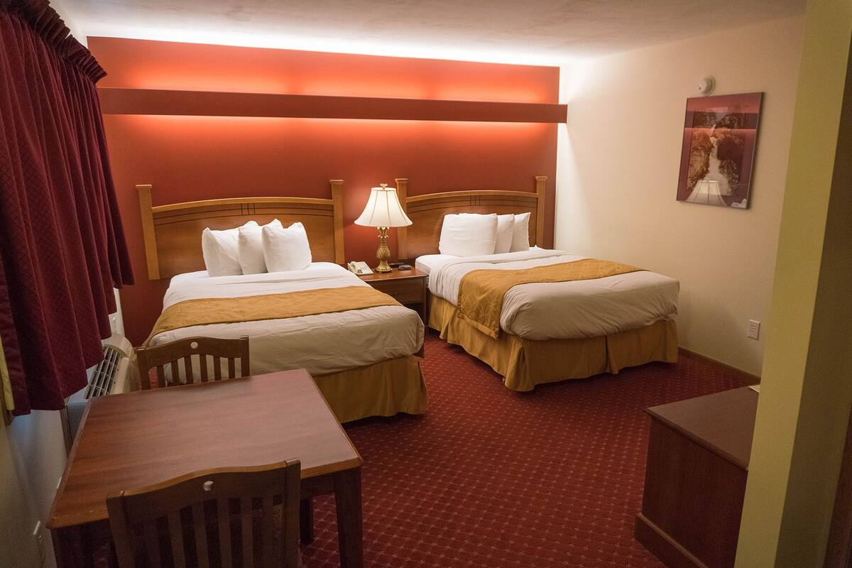 A hotel room with two beds, a desk, and a chair. There is a lamp on the desk and a painting on the wall.