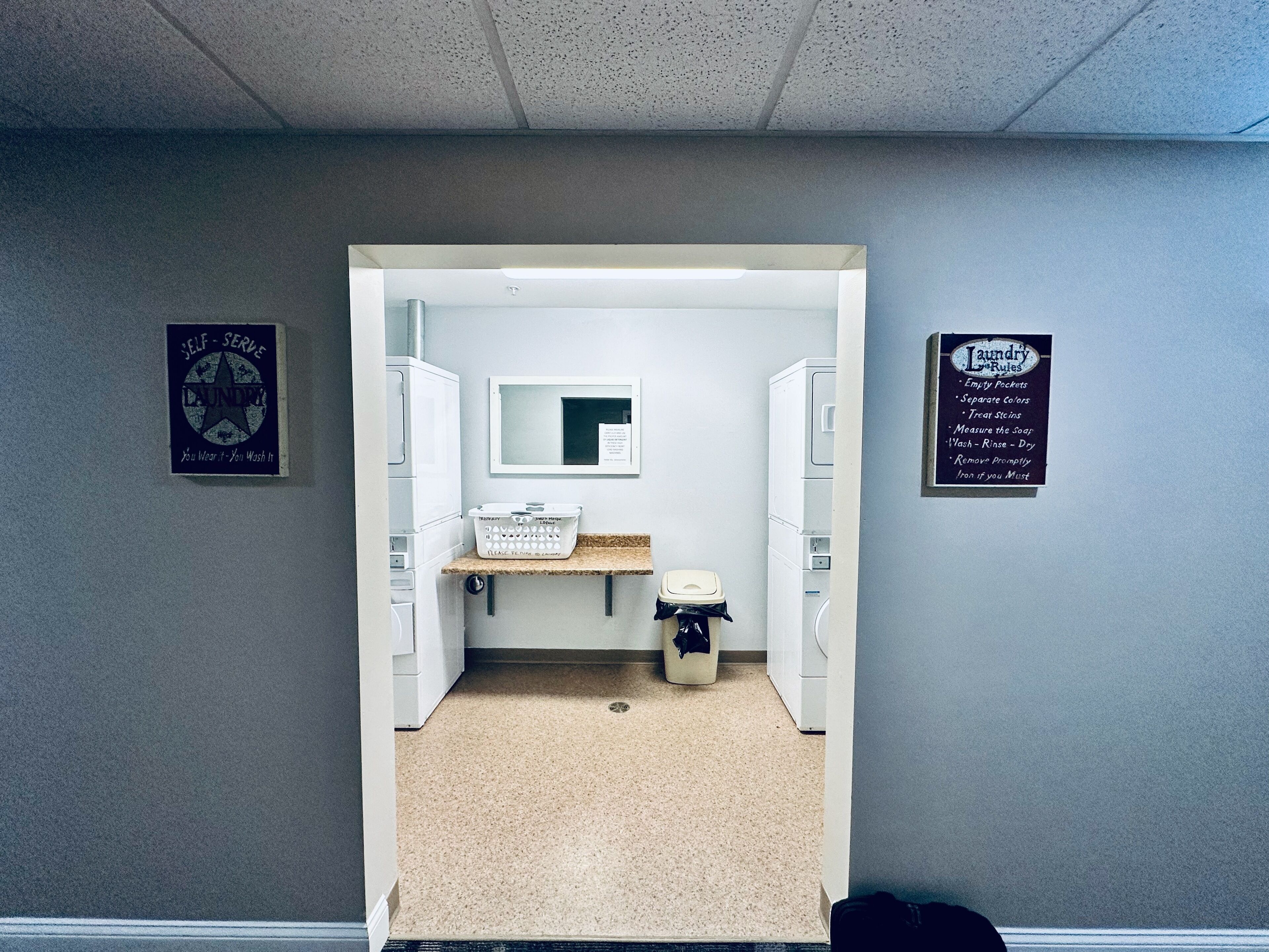 A small laundry room with a sink, a mirror, and a trash bin. There are two laundry baskets and a sign with laundry rules.