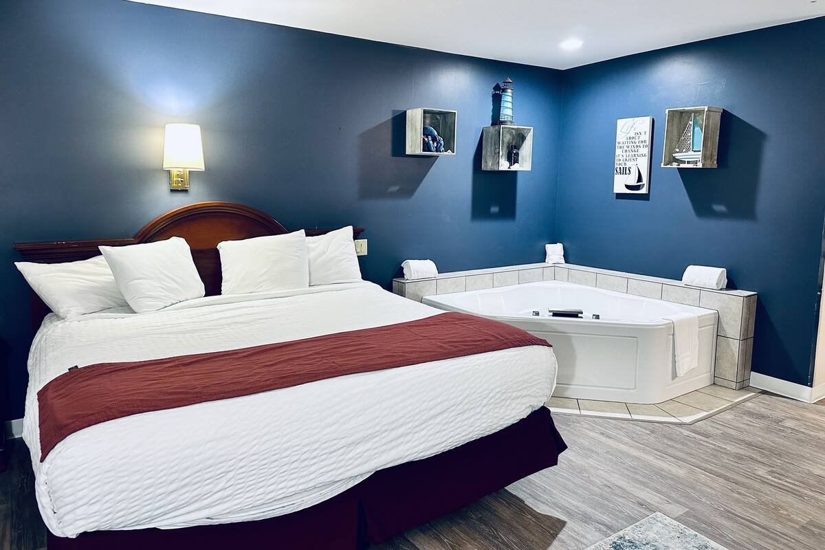 Habitación de hotel con una cama grande, una pequeña zona de estar y una bañera blanca, con una lámpara fijada a la pared.
