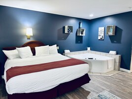Habitación de hotel con una cama grande, una pequeña zona de estar y una bañera blanca, con una lámpara fijada a la pared.