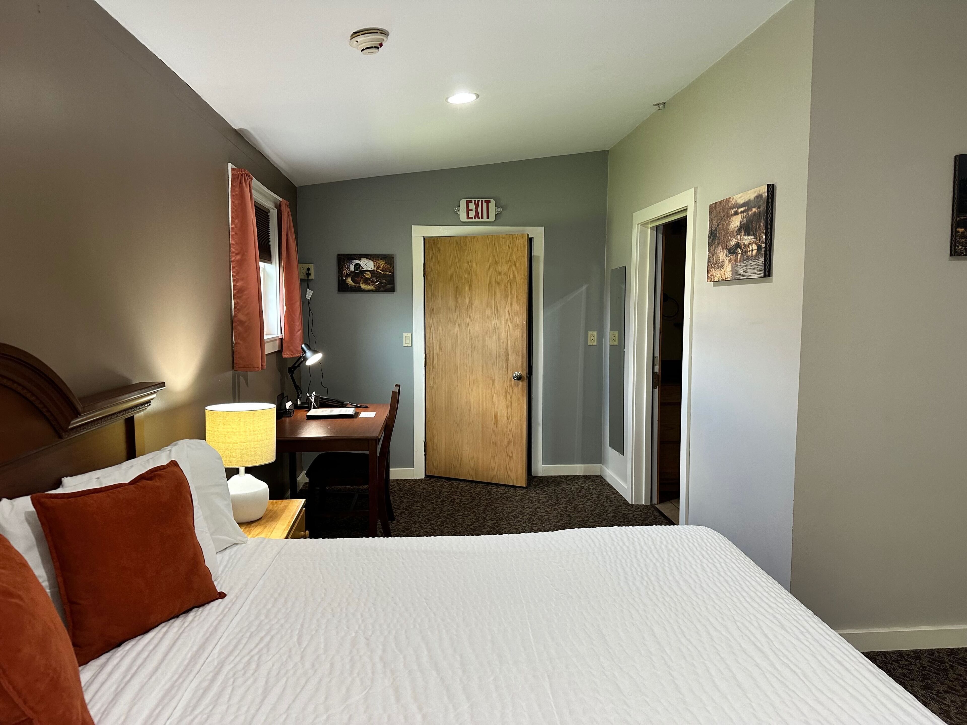 A hotel room with a bed, a desk, a lamp, an exit sign, and a door.