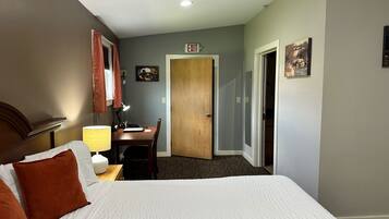 A hotel room with a bed, a desk, a lamp, an exit sign, and a door.