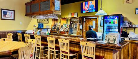 A bar area with wooden chairs, a bar counter, a television screen, and a Pepsi refrigerator.