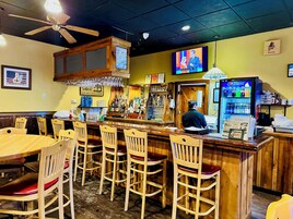 Una zona de bar con sillas de madera, una barra, una pantalla de televisión y un refrigerador de Pepsi.