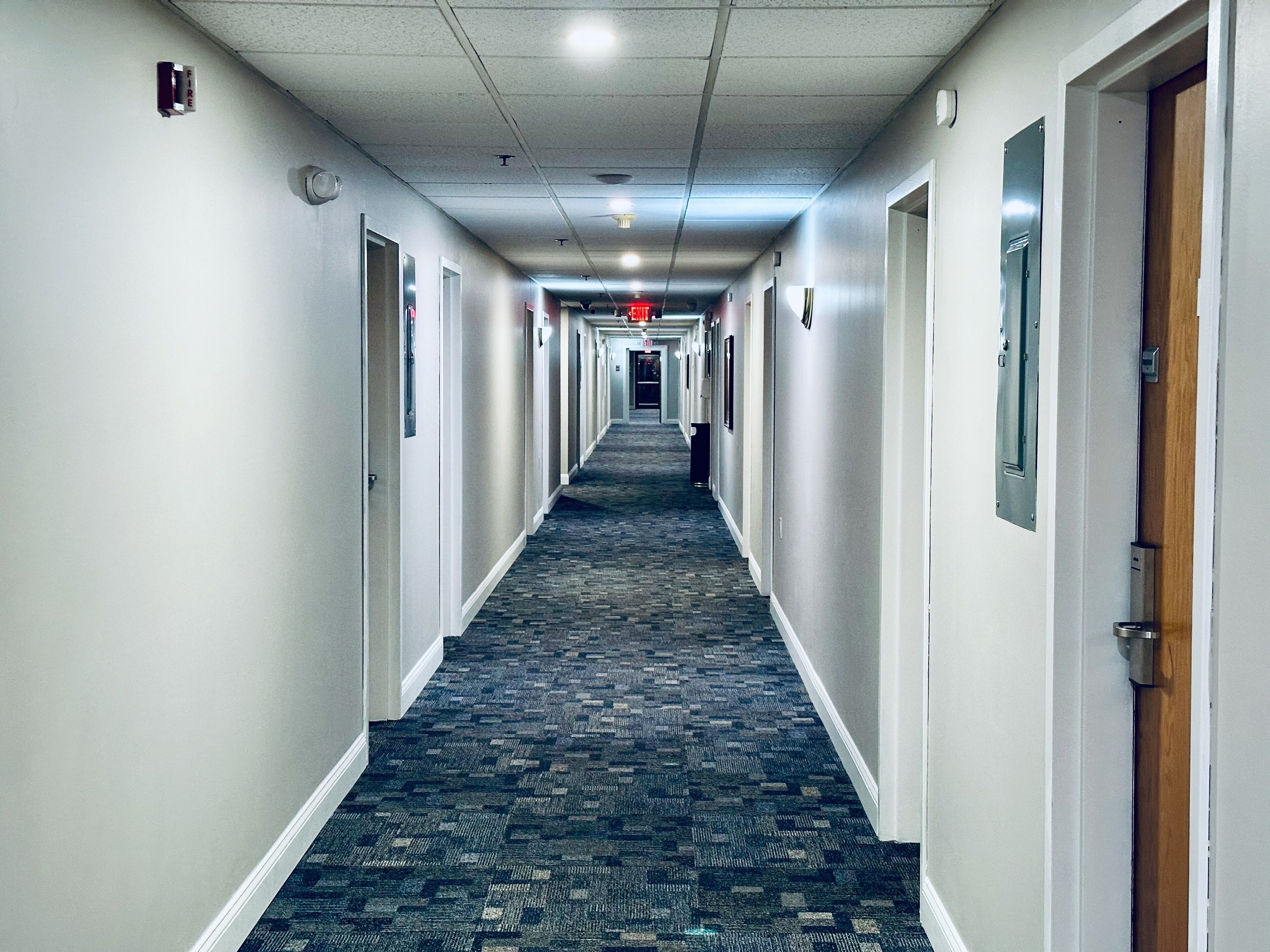 Un pasillo de hotel con pisos alfombrados, puertas a ambos lados y un detector de incendios en la pared izquierda.