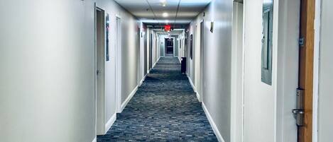 Un pasillo de hotel con pisos alfombrados, puertas a ambos lados y un detector de incendios en la pared izquierda.
