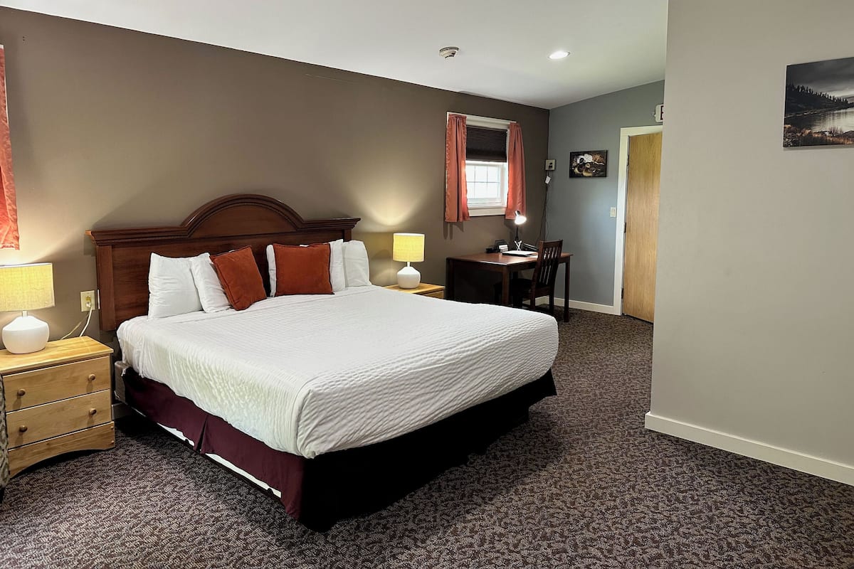 Un cuarto de hotel con una cama grande, mesitas de noche con lámparas, un escritorio con una silla, una ventana con cortinas y un cuadro en la pared.