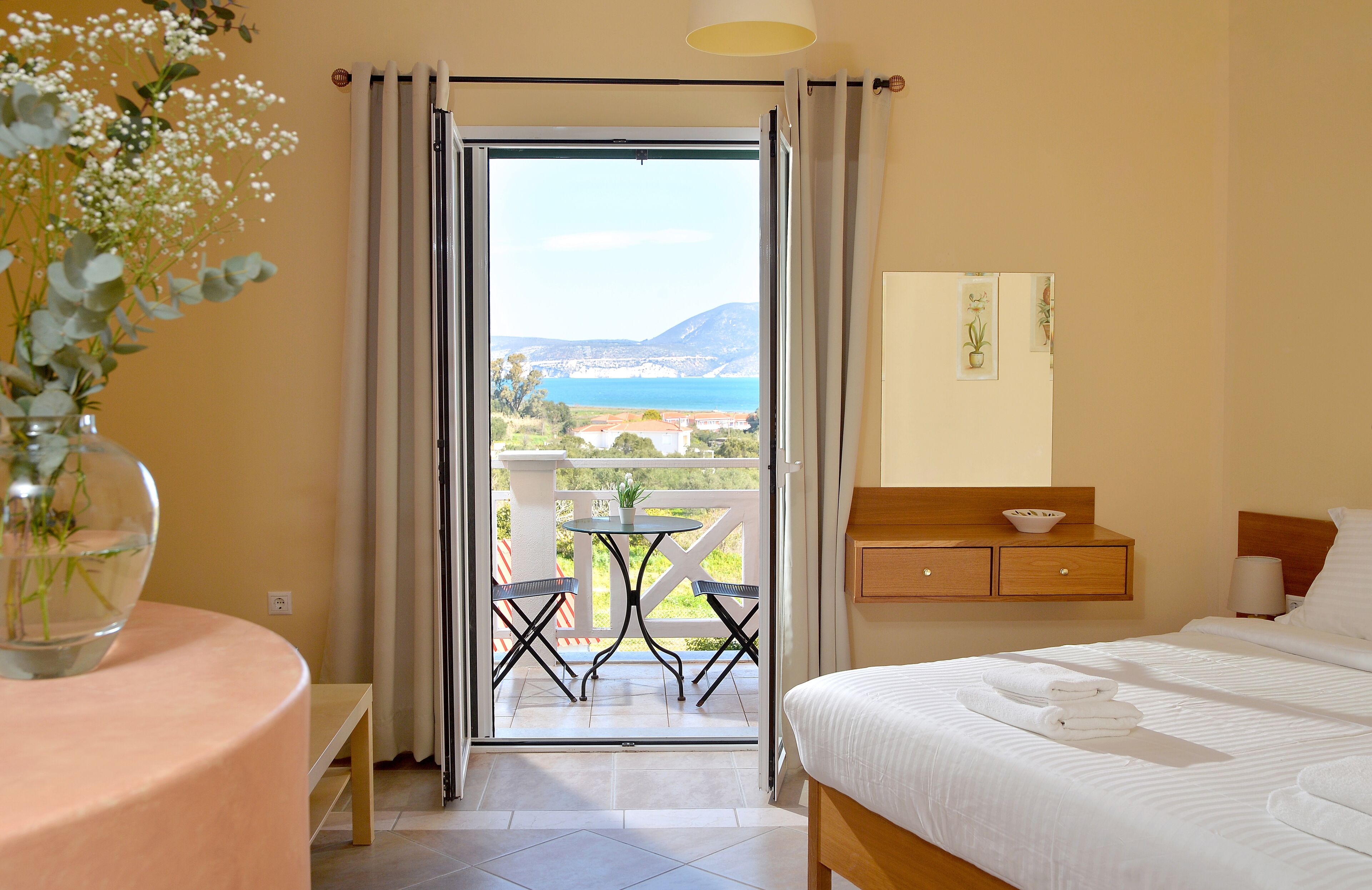 A hotel room with a bed, a bedside table, a small table with chairs, a view of the sea, and a balcony with a table and chairs.