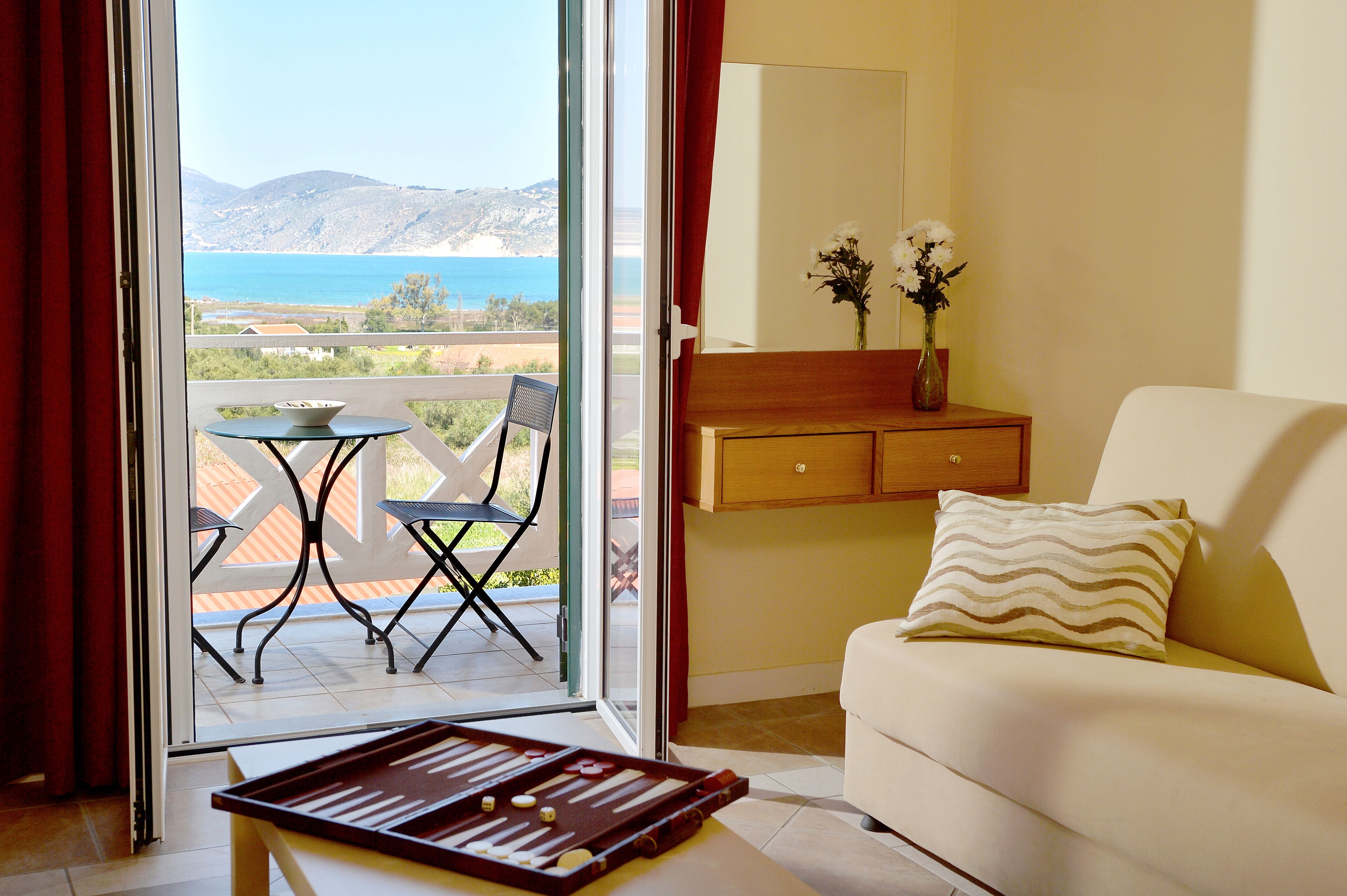 A room with a sofa, a small table, and a chair. There is a view of the sea and mountains through a glass door.