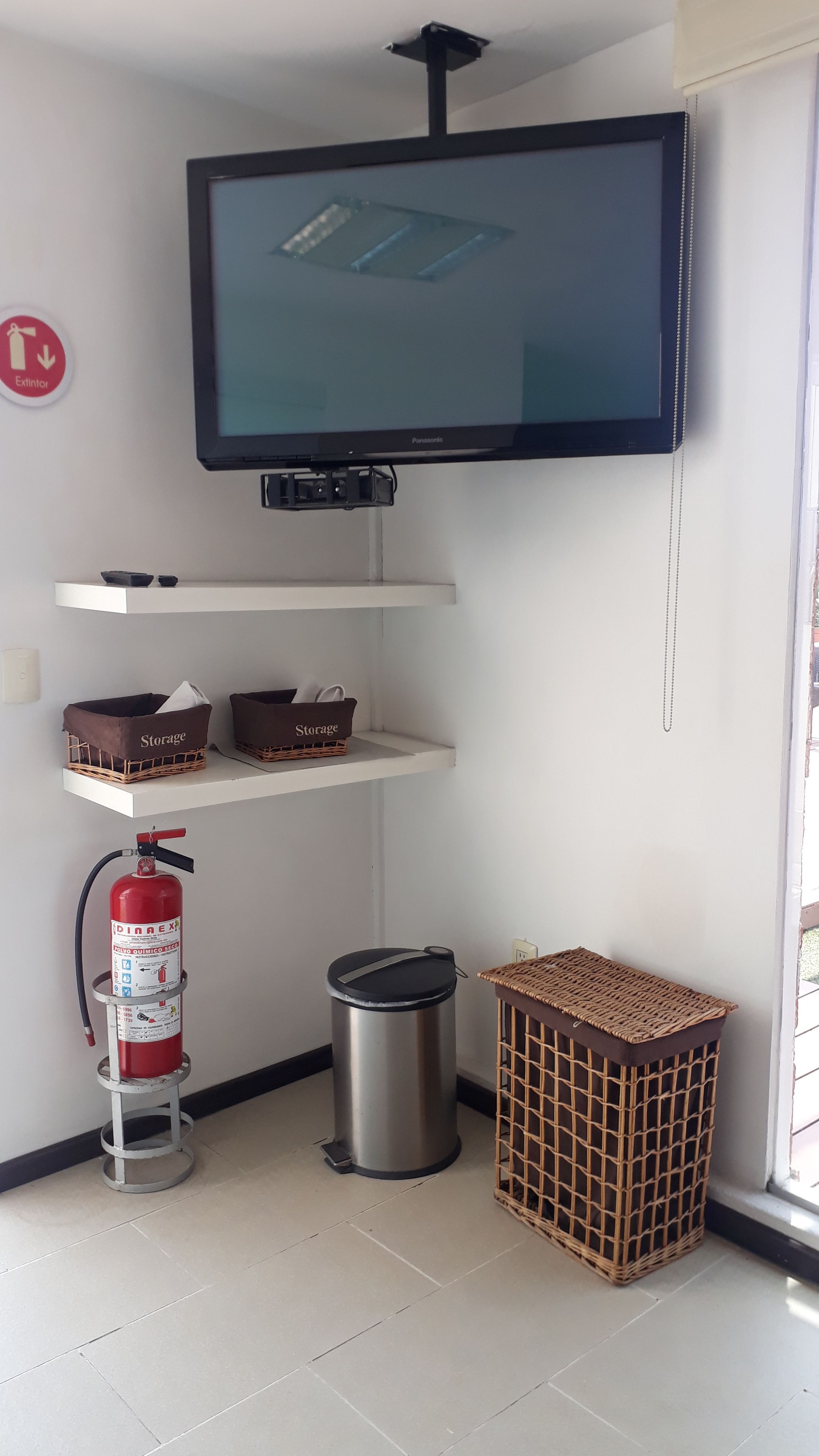Een kamer met een wandgemonteerde televisie, opbergmanden, een brandblusser, een prullenbak en een rieten mand.