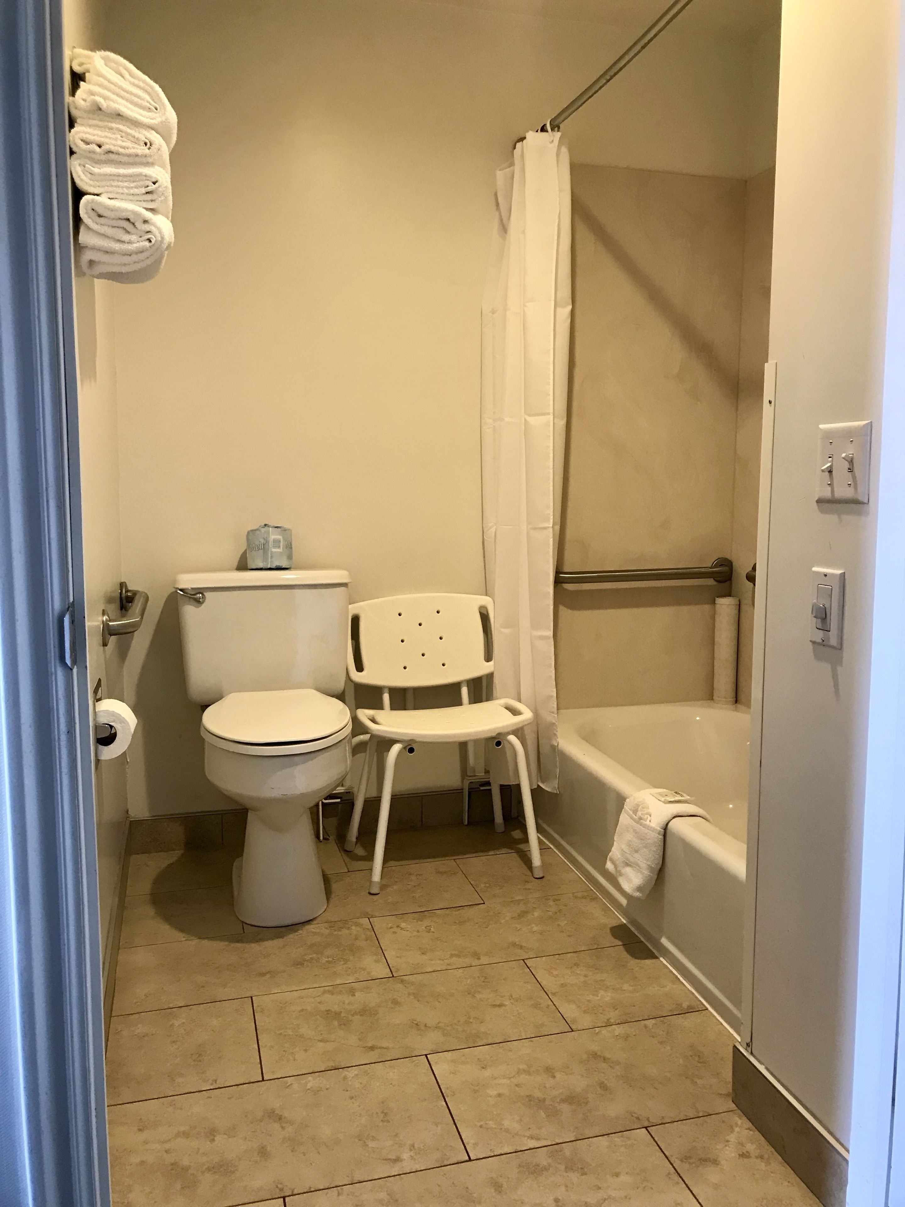 A bathroom with a toilet, a shower with a curtain, a bathtub, and a small white chair.