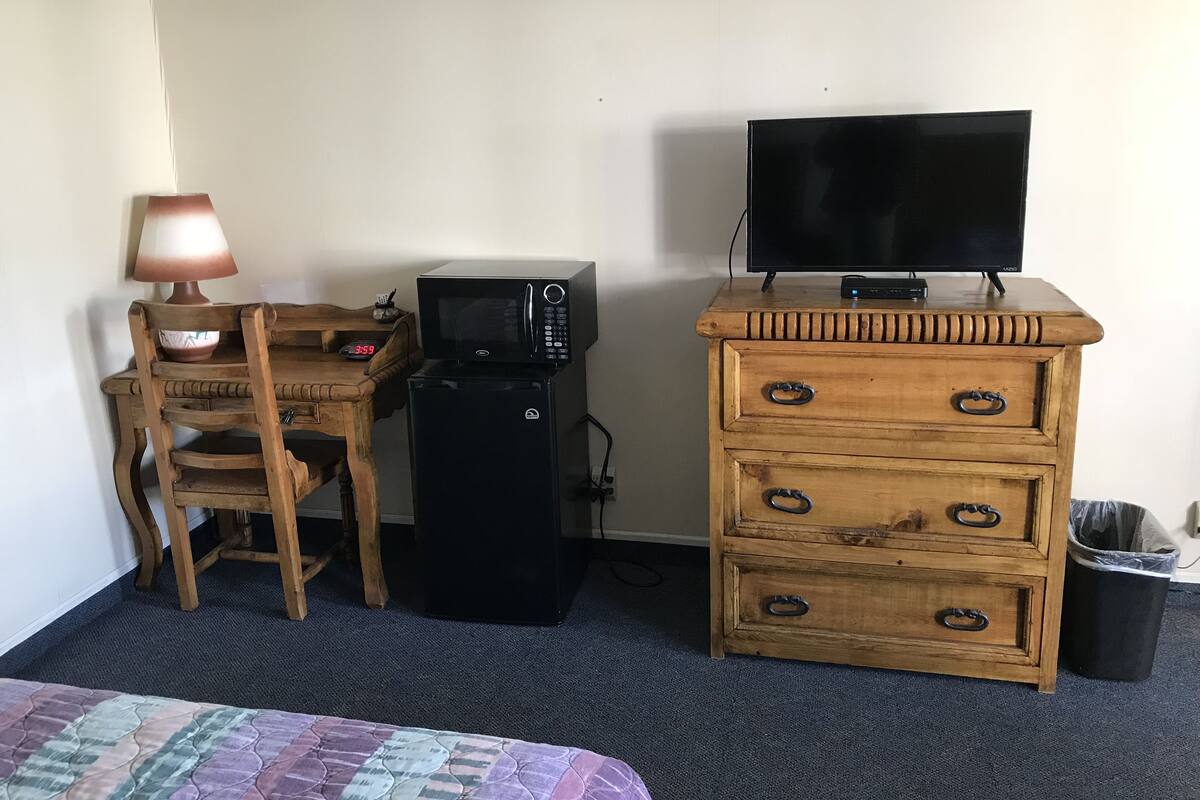 Une pièce comprenant un bureau en bois, un four à micro-ondes, un téléviseur posé sur une commode, une lampe et une poubelle.