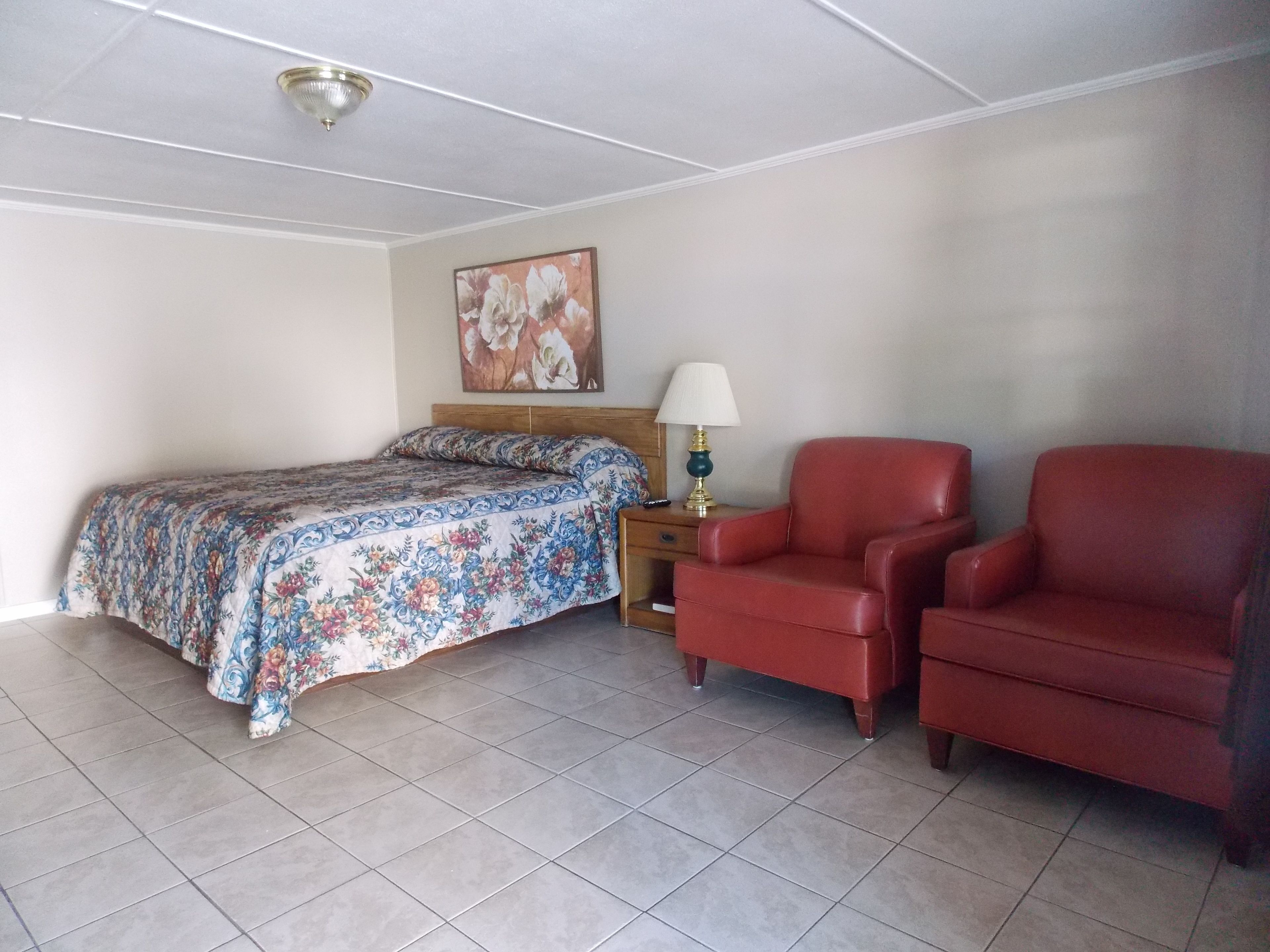 Une chambre à coucher avec un lit, deux fauteuils rouges, une table de chevet avec une lampe et un tableau floral accroché au mur.