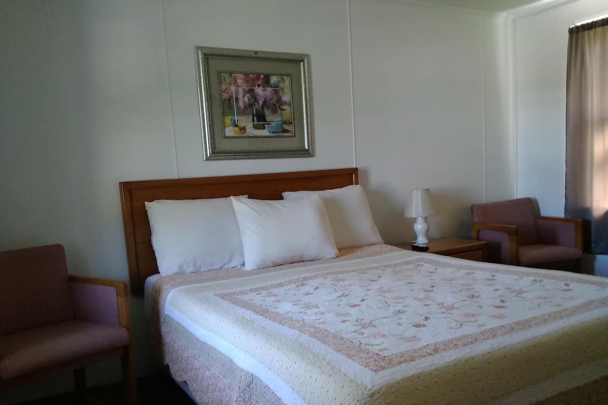 Quarto de hotel com uma cama grande, mesa de cabeceira, luminária, duas cadeiras e um quadro na parede.