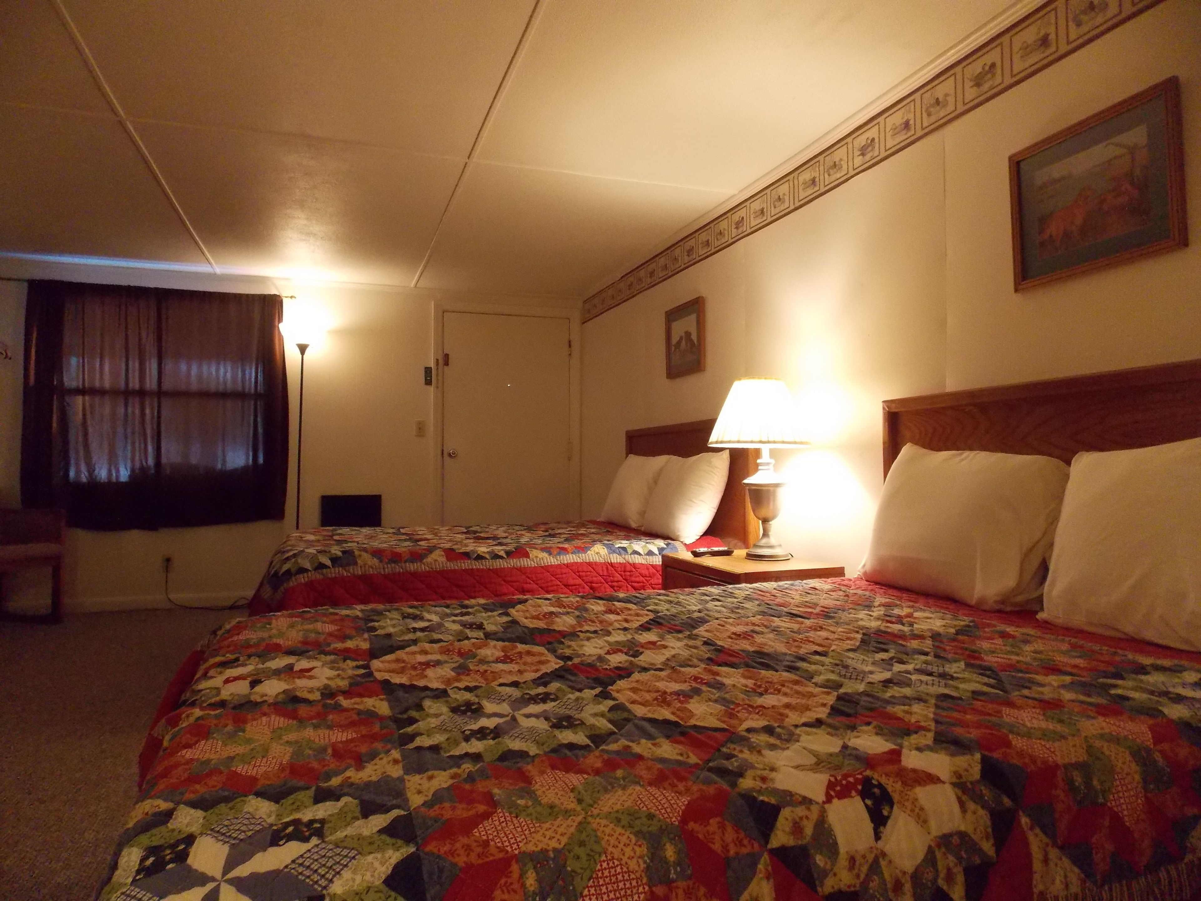 Une chambre d’hôtel avec deux lits, une table de chevet avec une lampe, un tableau au mur et une fenêtre avec des rideaux.