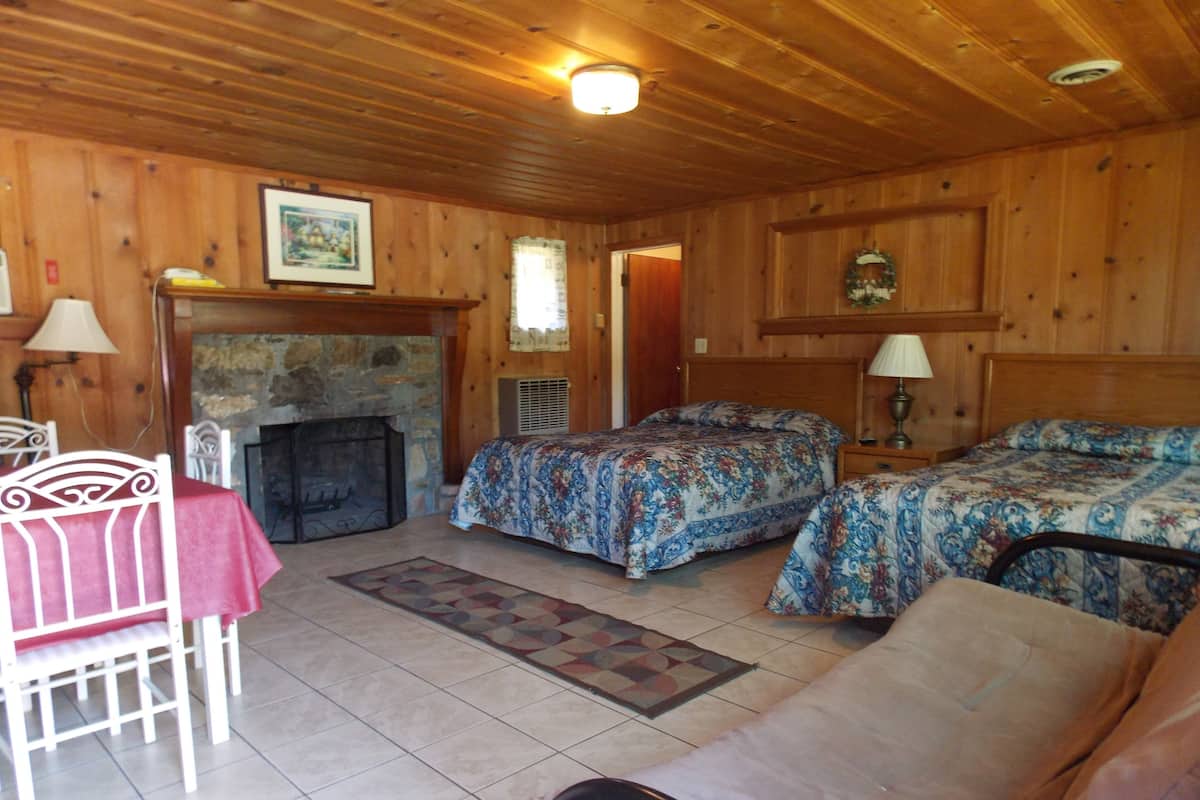 Um quarto acolhedor em estilo cabana, com lareira, duas camas com roupa de cama florida, uma mesa de jantar com toalha vermelha e teto de madeira.