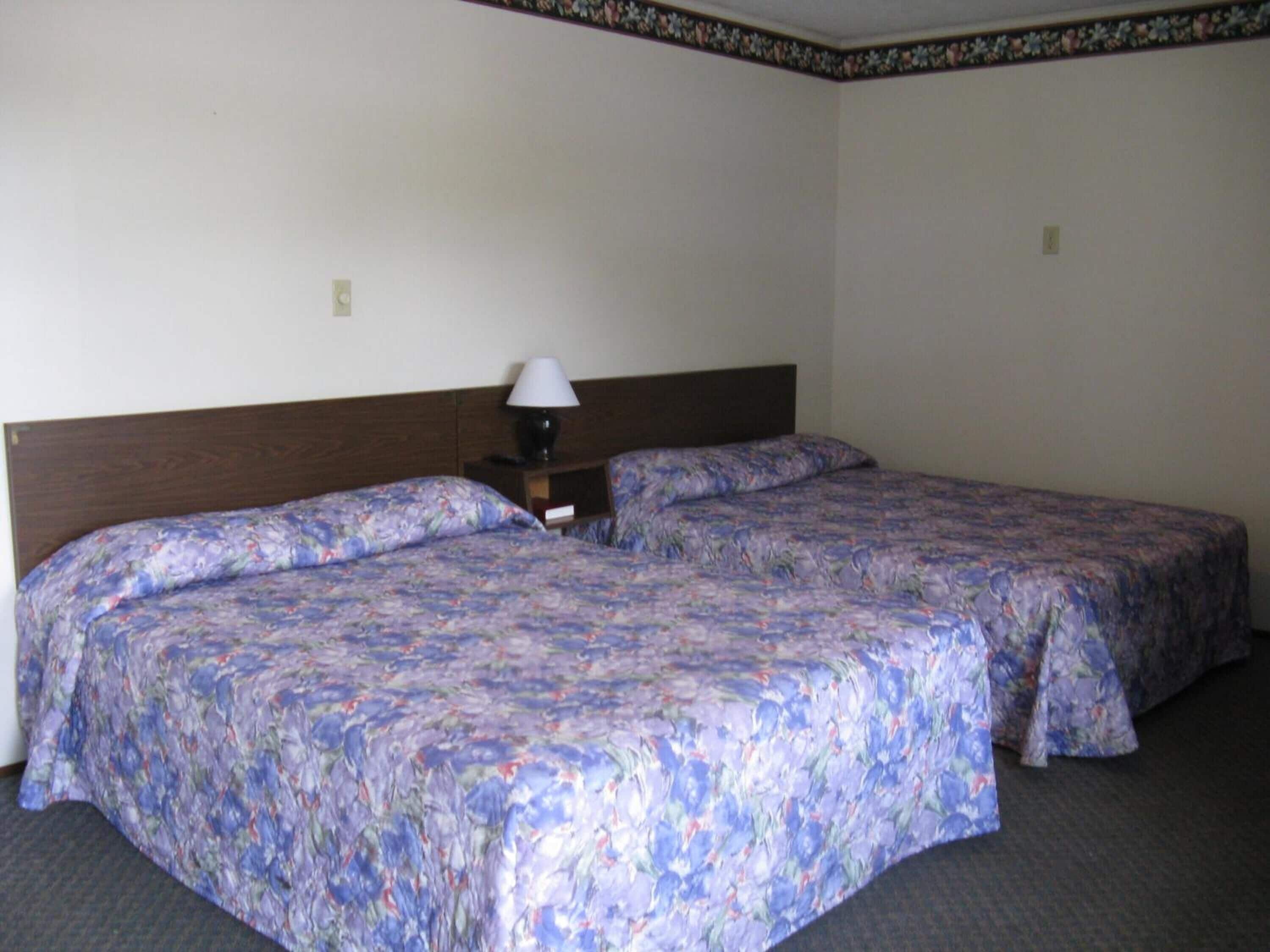 A hotel room with two single beds, a wooden headboard, a nightstand with a lamp, and a floral-patterned bedspread.