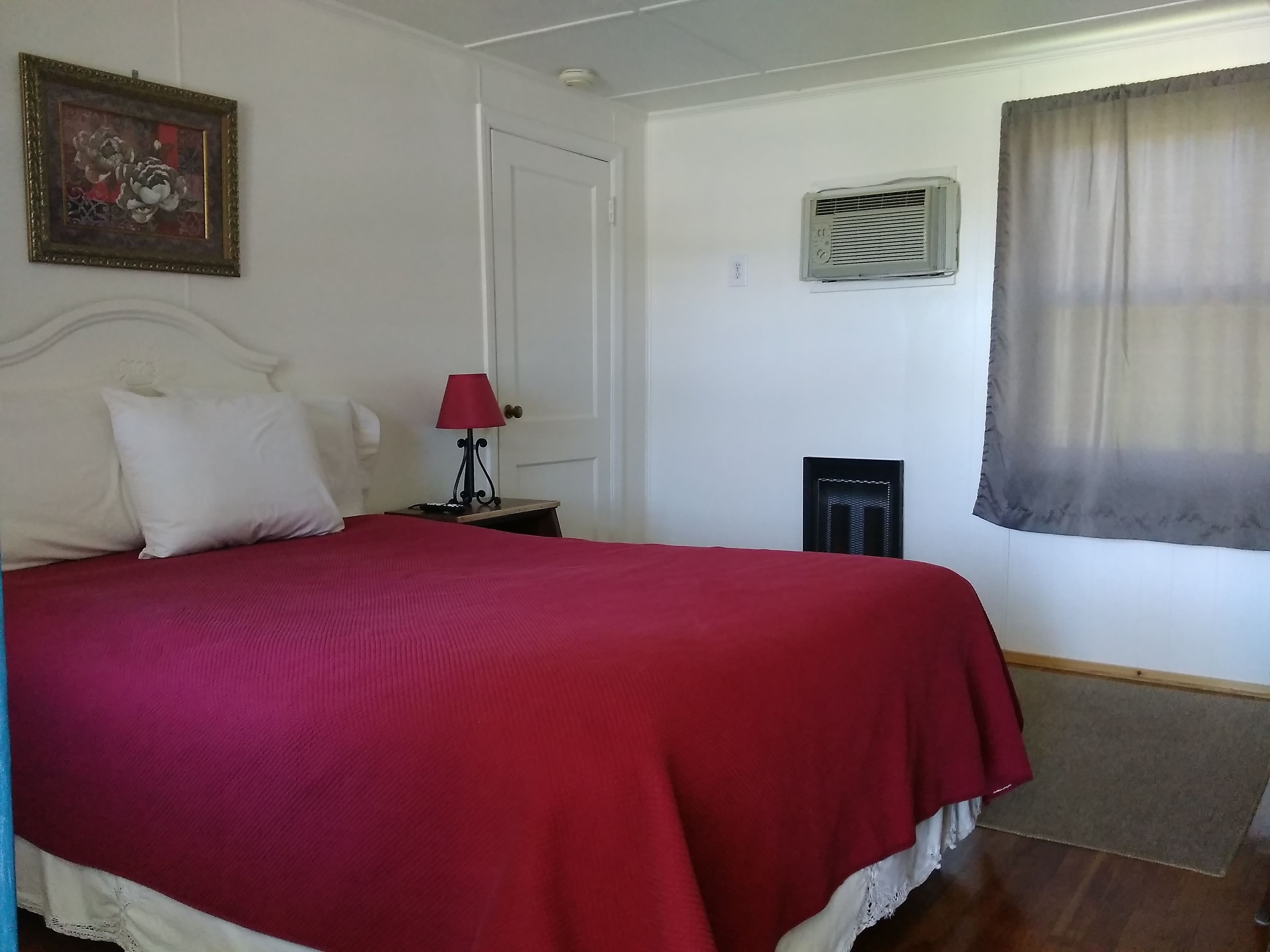 A bedroom with a large bed, a nightstand with a lamp, a framed picture on the wall, and an air conditioning unit.