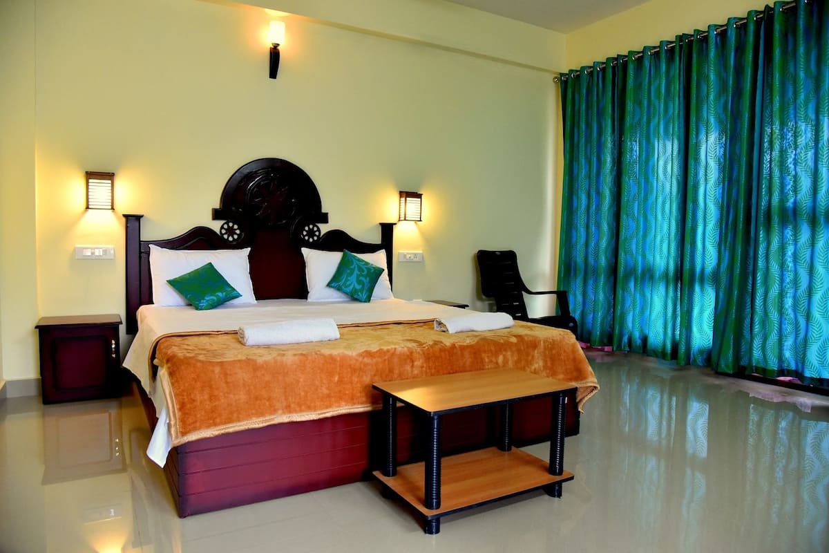 A hotel room with a bed, bedside lamps, a nightstand, a wooden bench, and a large window with curtains.