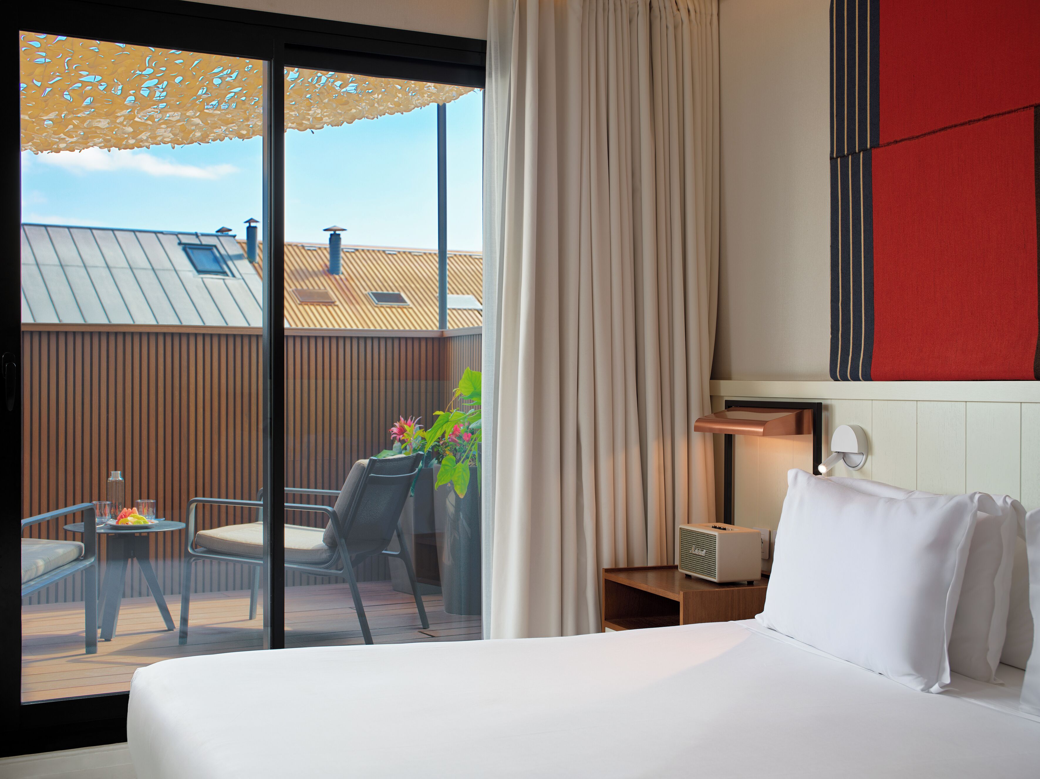 A hotel room with a bed, a small table with a lamp, a chair, and a view of the outside balcony.