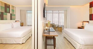 A hotel room with two beds, a desk with a lamp, a TV mounted on the wall, and a large window with curtains.