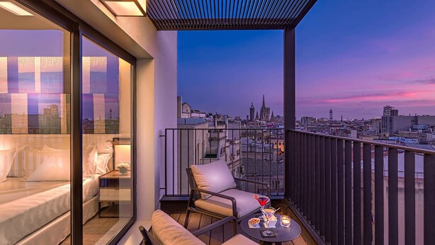 Balcón al atardecer con vista a la ciudad, que incluye un sofá, sillas y una pequeña mesa con aperitivos y una bebida.