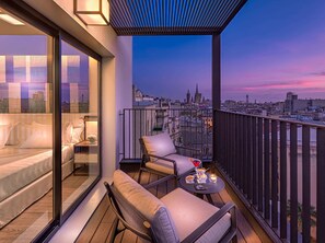 Balcón al atardecer con vista a la ciudad, que incluye un sofá, sillas y una pequeña mesa con aperitivos y una bebida.