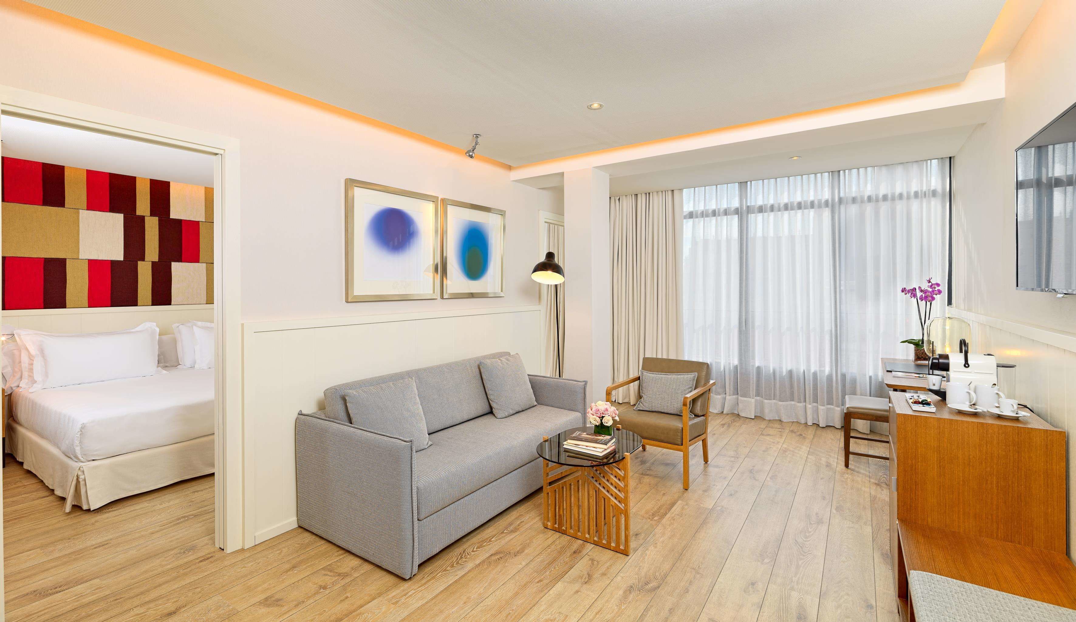 A modern hotel room with a grey sofa, a wooden coffee table, a small dining table with a chair, and a bed with white bedding.