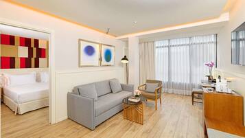 A modern hotel room with a grey sofa, a wooden coffee table, a small dining table with a chair, and a bed with white bedding.