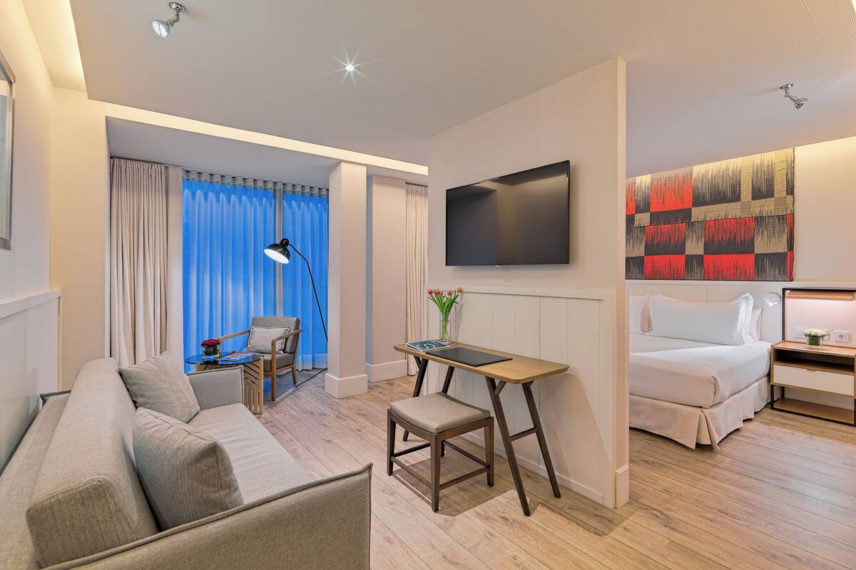 A modern hotel room with a sofa, a small table, a TV, a bed, and a large abstract painting on the wall.
