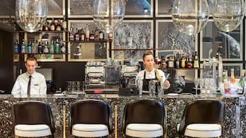 A bar area with a marble countertop, two baristas, a coffee machine, and a variety of bottles and glasses.
