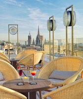 Een terras op het dak met rieten meubels, een tafel met een drankje en uitzicht op een stadshemelbeeld, waaronder een kathedraal.