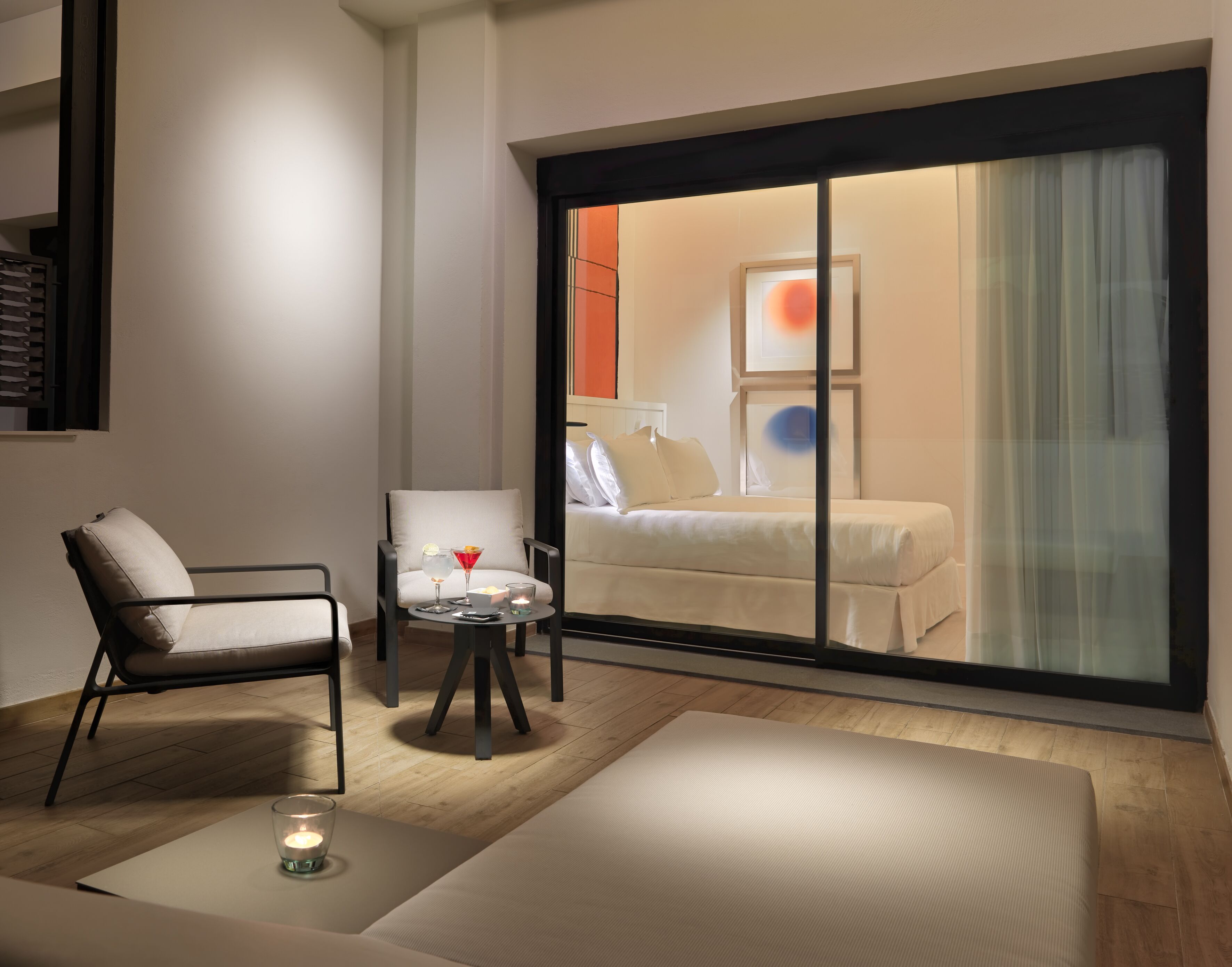 A modern hotel room with a large sliding glass door leading to a balcony, a sofa, a small table with a candle, and a chair.