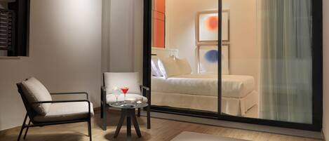 A modern hotel room with a large sliding glass door leading to a balcony, a sofa, a small table with a candle, and a chair.