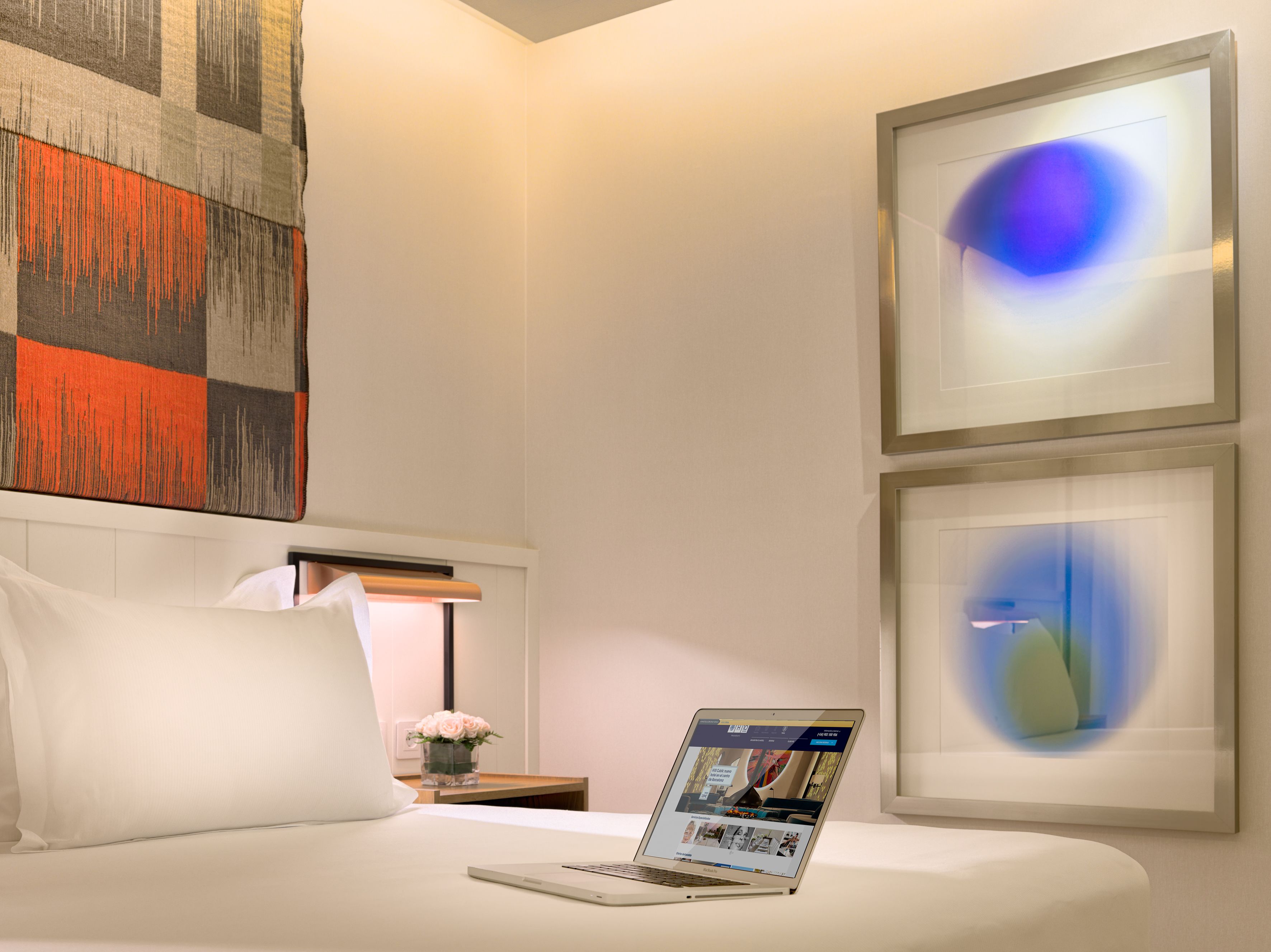 Une chambre d’hôtel avec un lit, un ordinateur portable posé sur le lit, une table de chevet ornée d’un vase de fleurs et deux tableaux abstraits accrochés au mur.