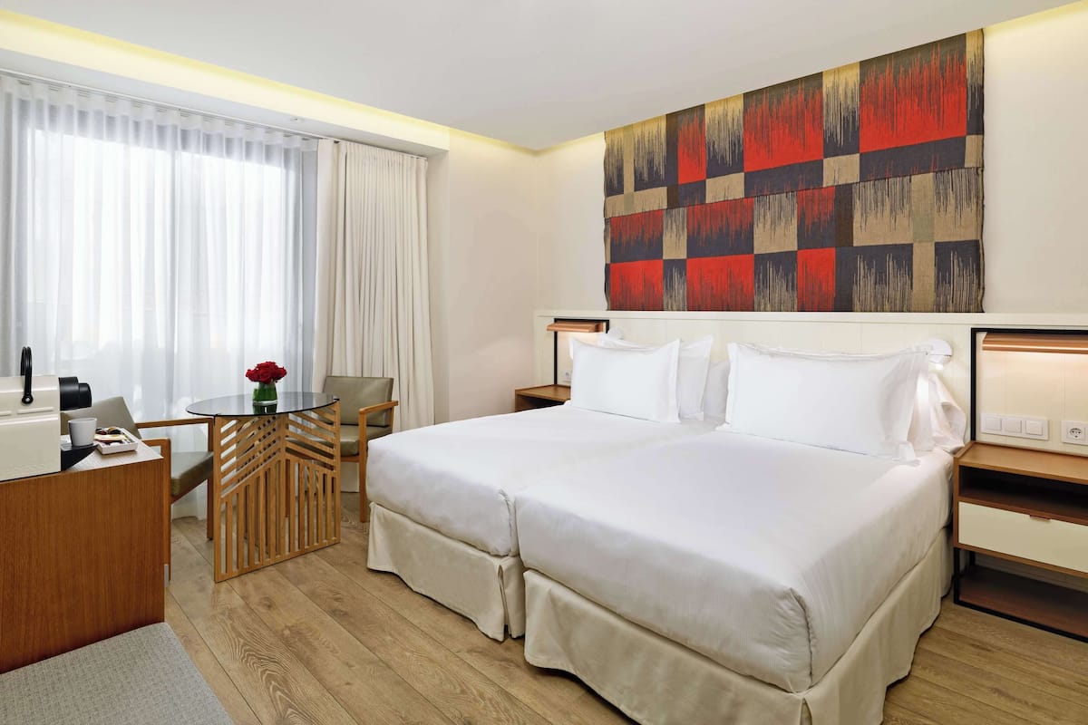 Ein Hotelzimmer mit Bett, Schreibtisch mit Kaffeemaschine, Stuhl, kleinem Tisch mit Blumenvase und großem abstrakten Gemälde an der Wand.