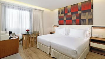 A hotel room with a bed, a desk with a coffee machine, a chair, a small table with a vase of flowers, and a large abstract painting on the wall.