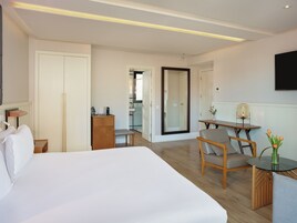 A hotel room with a large bed, a desk with a chair, a small table with a vase of flowers, a wardrobe, and a mirror.