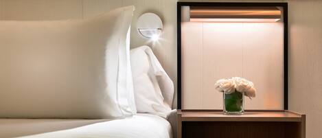 A neatly made bed with white linens, a bedside table with a vase of flowers, and a wall-mounted light fixture.