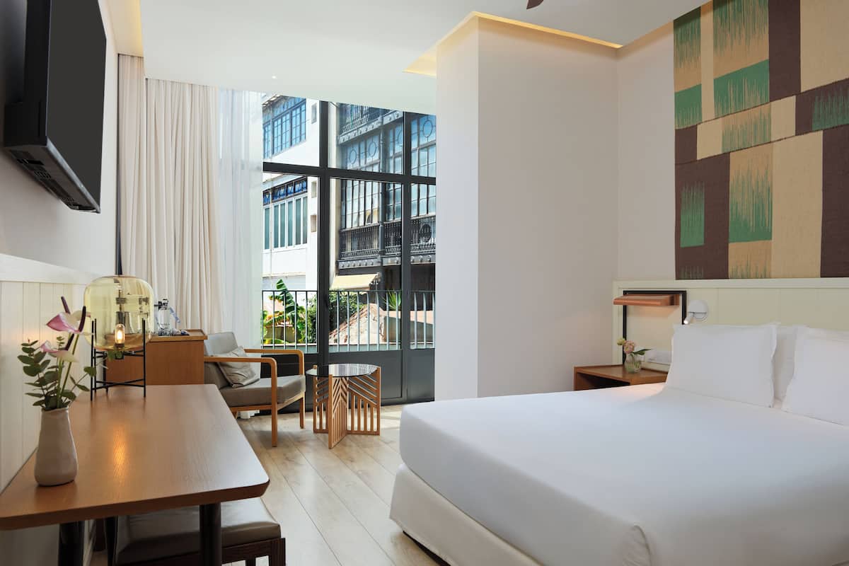 Ein modernes Hotelzimmer mit einem großen Bett, einem Schreibtisch mit Stuhl, einem kleinen Tisch mit Blumenvase und einem großen Fenster mit Blick auf Gebäude.