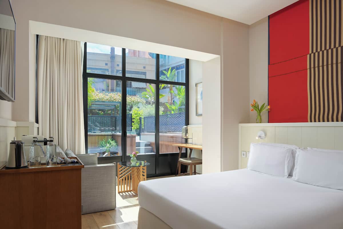 Ein Hotelzimmer mit einem großen Bett, einem Schreibtisch mit Stuhl, einem kleinen Tisch mit Vase und Blick ins Grüne.