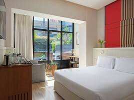 A hotel room with a large bed, a desk with a chair, a small table with a vase, and a view of a courtyard with plants and a building.
