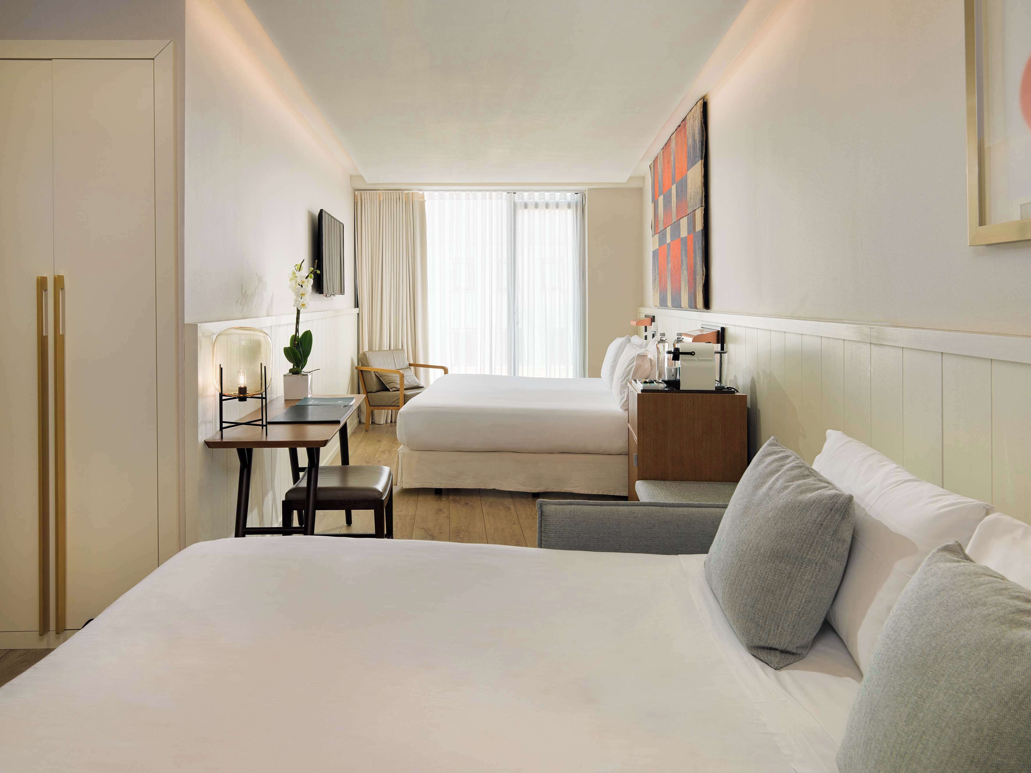 A hotel room with a large bed, a desk with a chair, a small table with a lamp, a TV mounted on the wall, and a window with curtains.