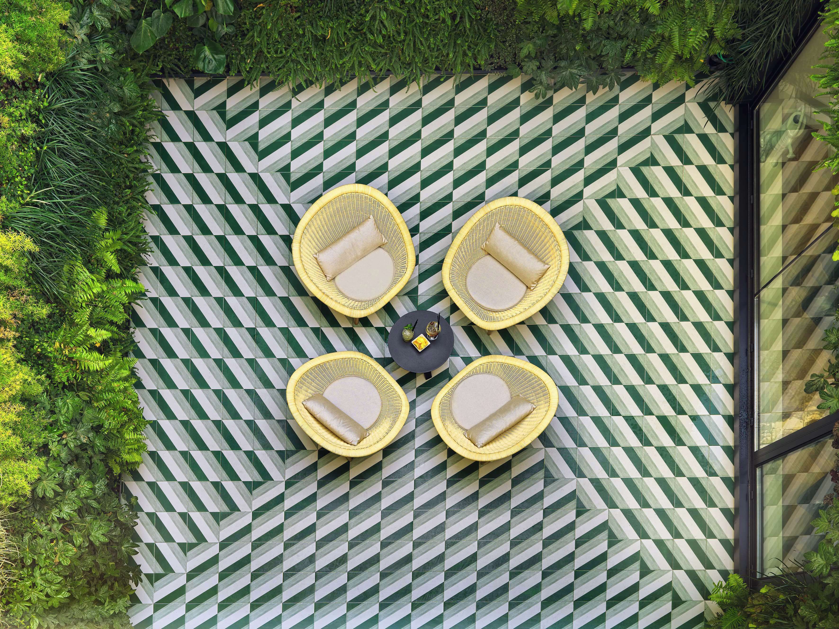 A top-down view of a patio area with a checkered floor pattern, four wicker chairs with cushions, and a small table with a bottle and glass.