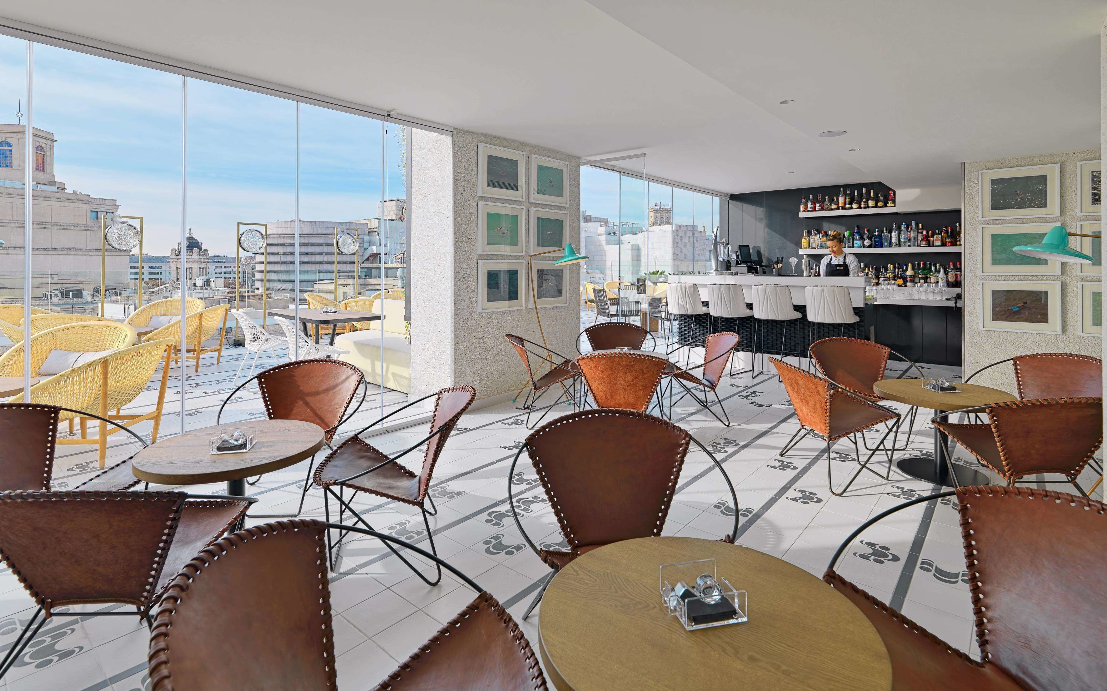 A rooftop bar with leather chairs and tables, a bartender, and a city view.