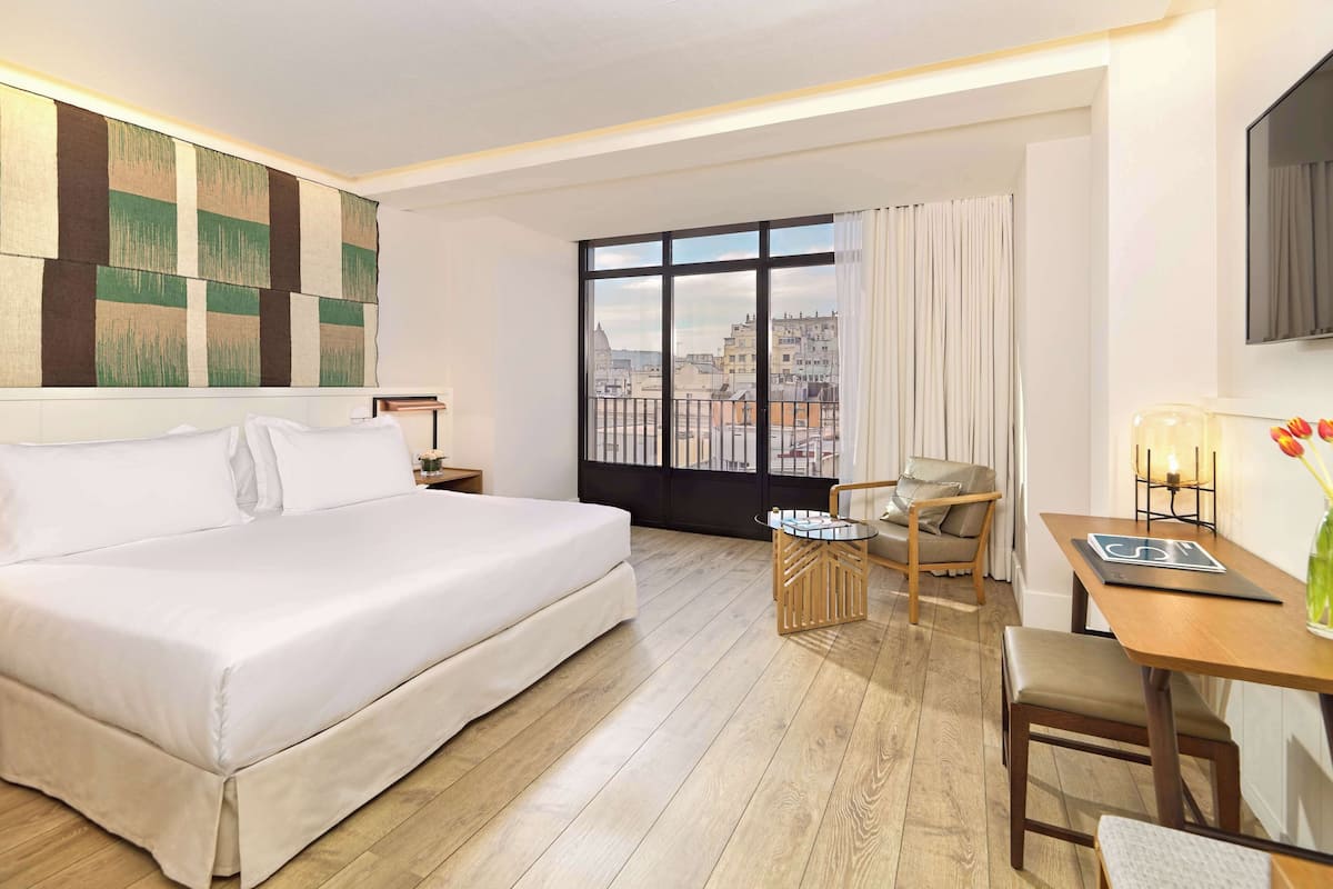 Ein Hotelzimmer mit einem gro ßen Bett, einem Schreibtisch, einem Sessel und Blick auf die Stadt durch ein großes Fenster.