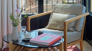 Un balcón con vistas a un paisaje urbano, una silla, una mesa con un libro, un florero con flores y un vaso de agua.